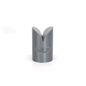 May include: Grey 3D printed object shaped like a cylinder with a split top and a stylised eye design on the front.