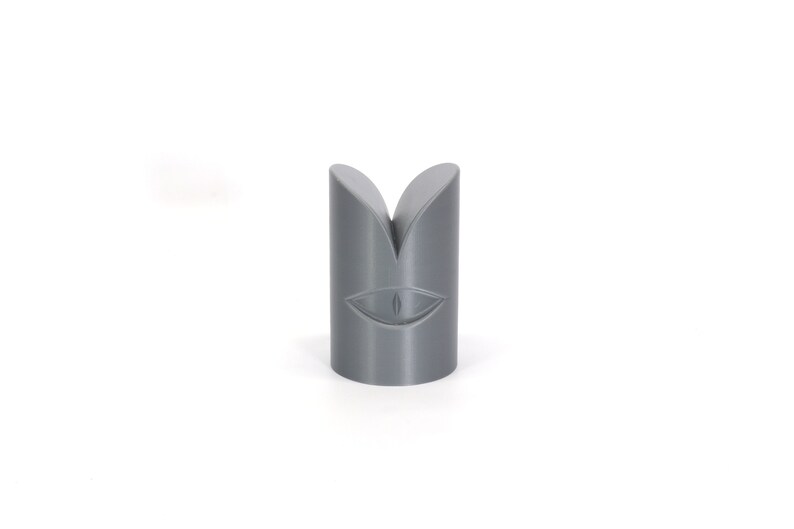 May include: Grey 3D printed object shaped like a cylinder with a split top and a stylised eye design on the front.