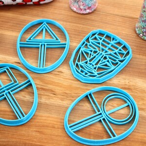 May include: Four blue plastic cookie cutters on a wooden surface. The cutters are shaped like a circle with a triangle inside, a circle with a square inside, a face, and a circle with a circle inside.