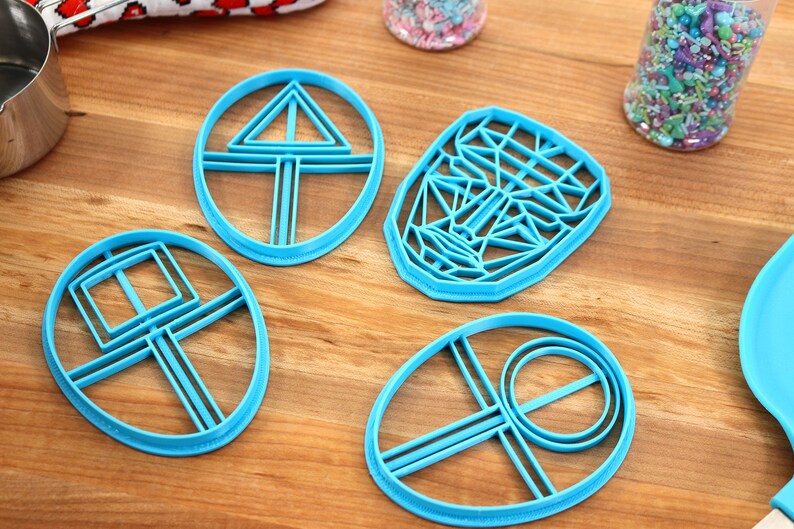 May include: Four blue plastic cookie cutters on a wooden surface. The cutters are shaped like a circle with a triangle inside, a circle with a square inside, a face, and a circle with a circle inside.