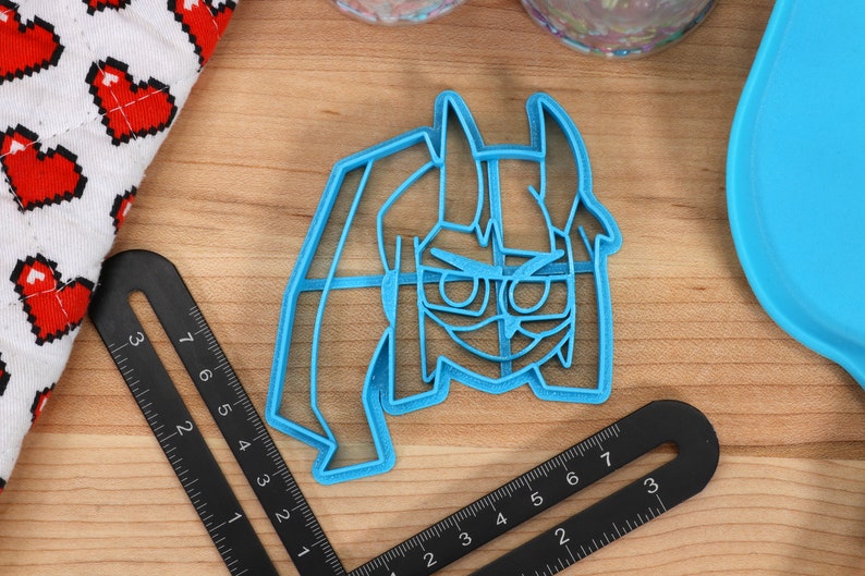 May include: A blue plastic cookie cutter shaped like a cartoon character with glasses and horns. The character has a serious expression.