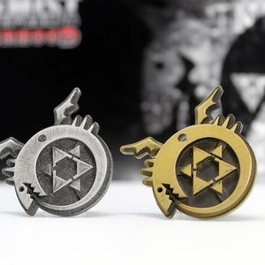 May include: Two metal pins with a black and gold finish. Each pin features a circular design with a stylized star inside. The star is surrounded by a circle with a jagged edge. The pins are on a white surface.