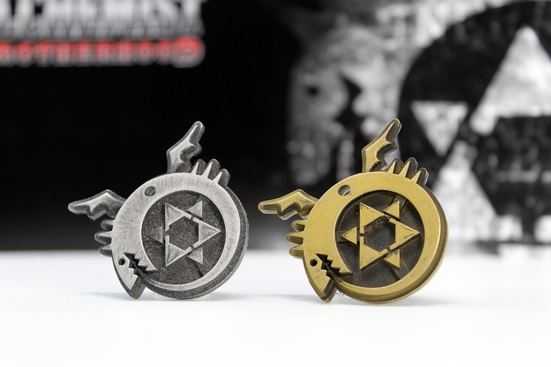 May include: Two metal pins with a black and gold finish. Each pin features a circular design with a stylized star inside. The star is surrounded by a circle with a jagged edge. The pins are on a white surface.