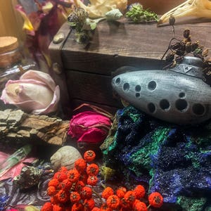 Puede incluir: Una ocarina plateada con un dise&ntilde;o triangular y m&uacute;ltiples agujeros, descansando sobre una roca azul y verde con bayas rojas en primer plano.