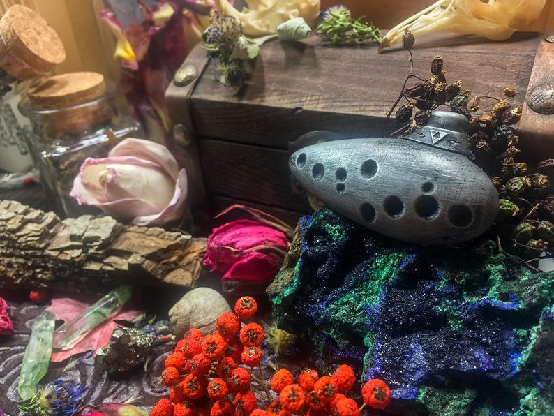 Puede incluir: Una ocarina plateada con un dise&ntilde;o triangular y m&uacute;ltiples agujeros, descansando sobre una roca azul y verde con bayas rojas en primer plano.