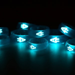May include: A group of eight glow-in-the-dark silicone rings with a white arrow design. The rings are stacked and arranged in a semi-circle on a black surface.