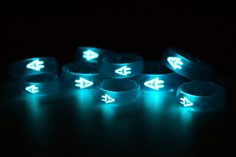 May include: A group of eight glow-in-the-dark silicone rings with a white arrow design. The rings are stacked and arranged in a semi-circle on a black surface.