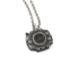 Puede incluir: Un collar plateado con un colgante con forma de dispositivo Digimon. El colgante tiene un dise&ntilde;o circular con una ventana cuadrada en el centro.