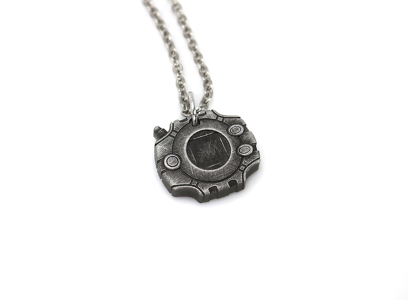 Puede incluir: Un collar plateado con un colgante con forma de dispositivo Digimon. El colgante tiene un dise&ntilde;o circular con una ventana cuadrada en el centro.