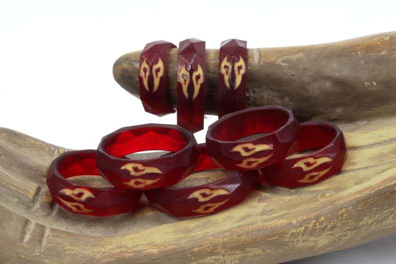 May include: A set of eight red resin rings with a geometric design and a yellow flame pattern. The rings are stacked on a wooden surface.