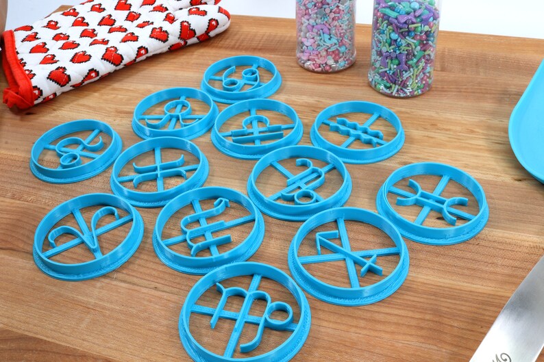 May include: A set of twelve blue plastic cookie cutters shaped like the astrological signs. The cutters are arranged on a wooden surface.