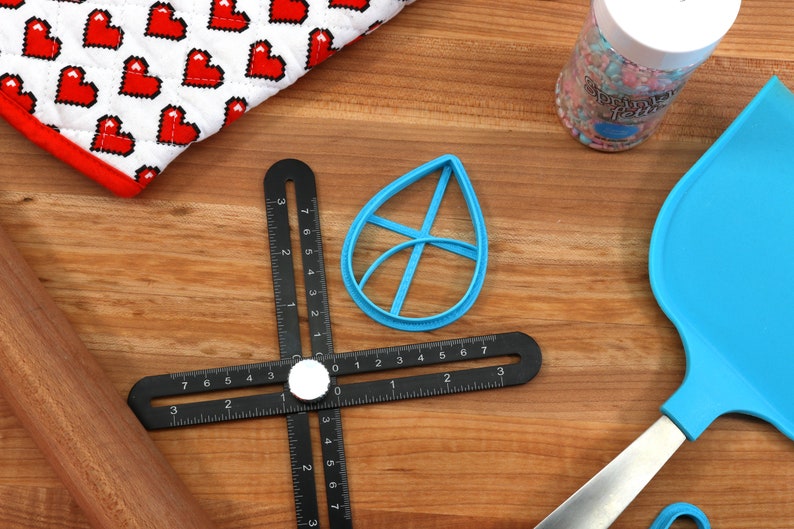 May include: A blue leaf-shaped cookie cutter, a black ruler with a rotating dial, a wooden rolling pin, a blue spatula, and a jar of colorful sprinkles on a wooden surface.
