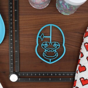 May include: A blue plastic cookie cutter shaped like a gorilla's face. The gorilla has a lightning bolt on its forehead and a white stripe across its nose.