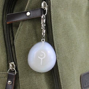 May include: A white, round keychain with a silver metal ring and a silver metal clasp. The keychain has a white symbol on it, which looks like a crescent moon and a staff.