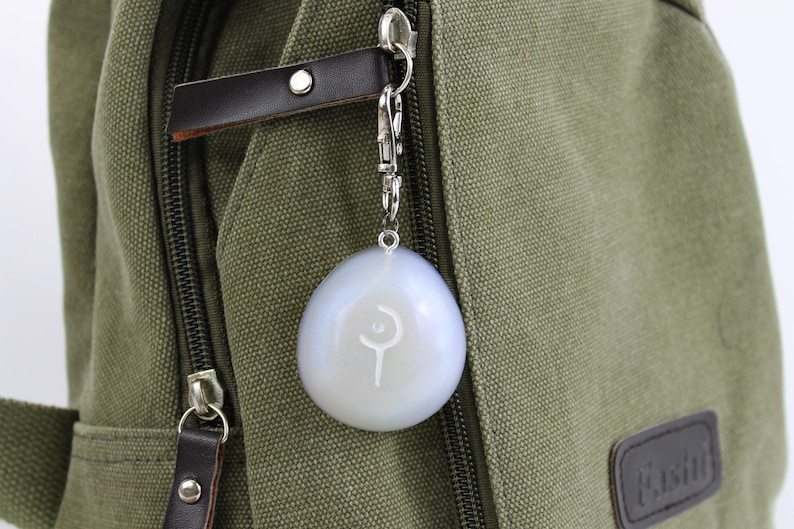 May include: A white, round keychain with a silver metal ring and a silver metal clasp. The keychain has a white symbol on it, which looks like a crescent moon and a staff.