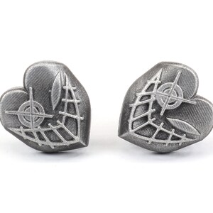 May include: A pair of silver heart-shaped earrings with a detailed design of a web and a crosshair.