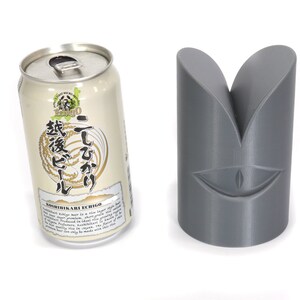 May include: A silver can of beer with Japanese text on the label. The text reads "Koshuhikari Echigo" and "Itegaki". The can is next to a grey, 3D printed object shaped like a face with a single eye.