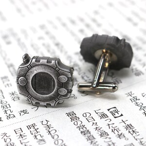 May include: A pair of silver cufflinks featuring a detailed design of a video game character. The cufflinks are on a white background.