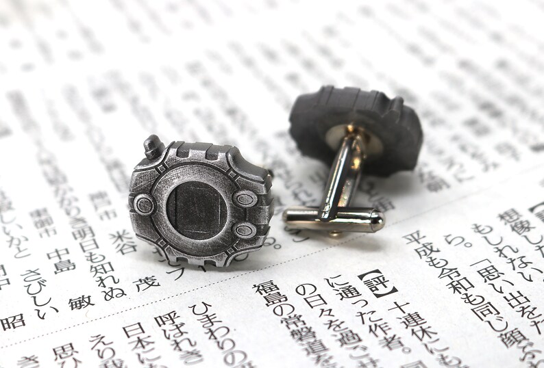 May include: A pair of silver cufflinks featuring a detailed design of a video game character. The cufflinks are on a white background.