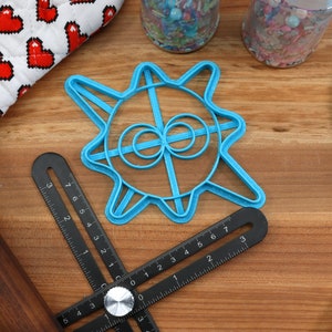 May include: A blue plastic cookie cutter shaped like a sun with a circle design in the center. The cutter is on a wooden surface with a rolling pin, a ruler, and a spatula.