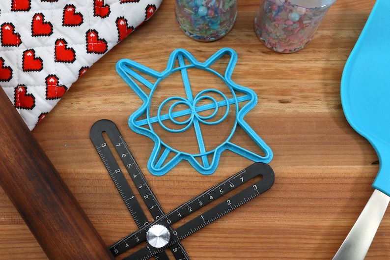 May include: A blue plastic cookie cutter shaped like a sun with a circle design in the center. The cutter is on a wooden surface with a rolling pin, a ruler, and a spatula.