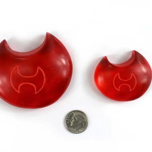 May include: Two red plastic musical instruments shaped like a crescent moon with a stylized axe symbol in the center. The larger instrument is about 4 inches in diameter and the smaller instrument is about 2 inches in diameter.