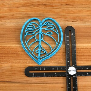 May include: A blue plastic cookie cutter shaped like a Monstera leaf, measuring approximately 5 inches in length, is placed on a wooden surface next to a black measuring tool.