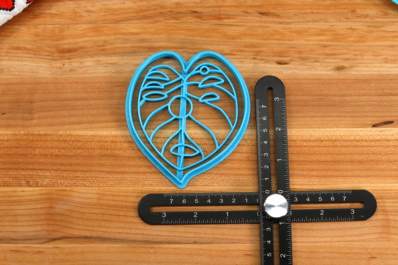 May include: A blue plastic cookie cutter shaped like a Monstera leaf, measuring approximately 5 inches in length, is placed on a wooden surface next to a black measuring tool.
