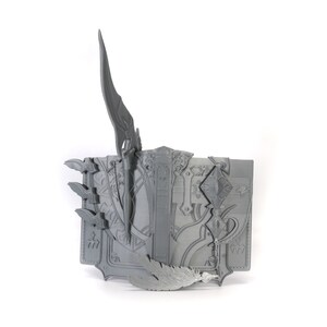 May include: A gray 3D printed book with intricate designs and a sword sticking out of the top. The book is open and has a feather sticking out of the side.