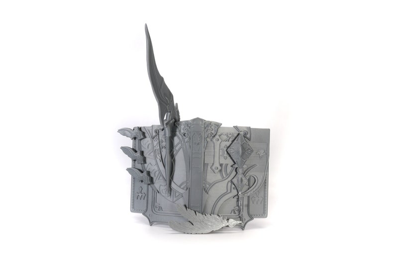May include: A gray 3D printed book with intricate designs and a sword sticking out of the top. The book is open and has a feather sticking out of the side.