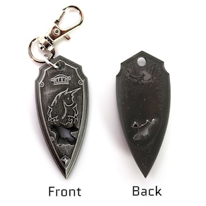 May include: A silver-colored keychain shaped like a shield with a unicorn design on the front and a plain back. The keychain has a silver-colored metal loop.