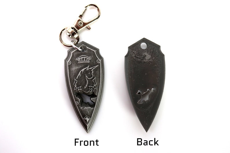 May include: A silver-colored keychain shaped like a shield with a unicorn design on the front and a plain back. The keychain has a silver-colored metal loop.