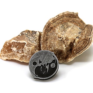 May include: A black and silver yin yang symbol pendant with a textured surface. The pendant is resting on two pieces of brown and white rock.