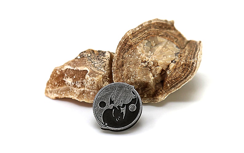 May include: A black and silver yin yang symbol pendant with a textured surface. The pendant is resting on two pieces of brown and white rock.