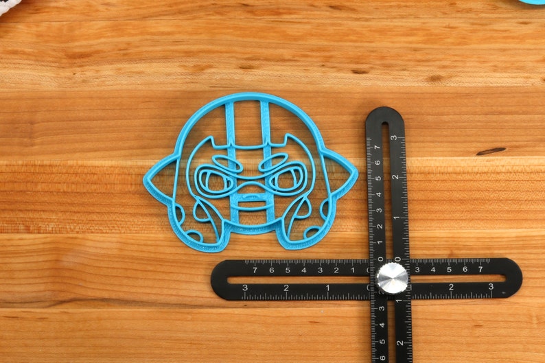 May include: A blue plastic cookie cutter shaped like a cartoon character with a helmet and a serious expression. The cutter is about 2 inches wide and is sitting on a wooden surface next to a ruler.