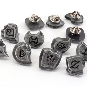 May include: A set of ten silver-toned metal pins featuring various symbols and designs. The pins are shaped like shields and have a textured surface.