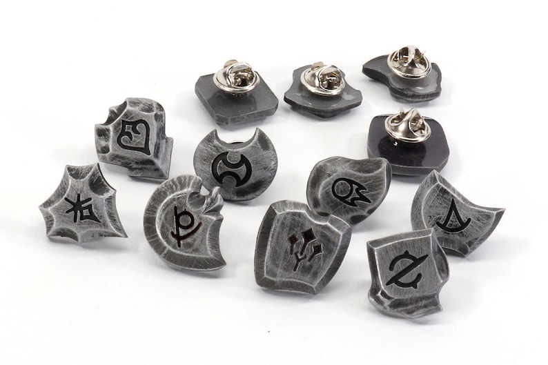 May include: A set of ten silver-toned metal pins featuring various symbols and designs. The pins are shaped like shields and have a textured surface.