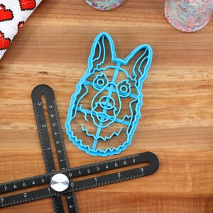 May include: A blue plastic cookie cutter shaped like a German Shepherd dog's head. The cutter is on a wooden surface with a ruler and a metal spatula.