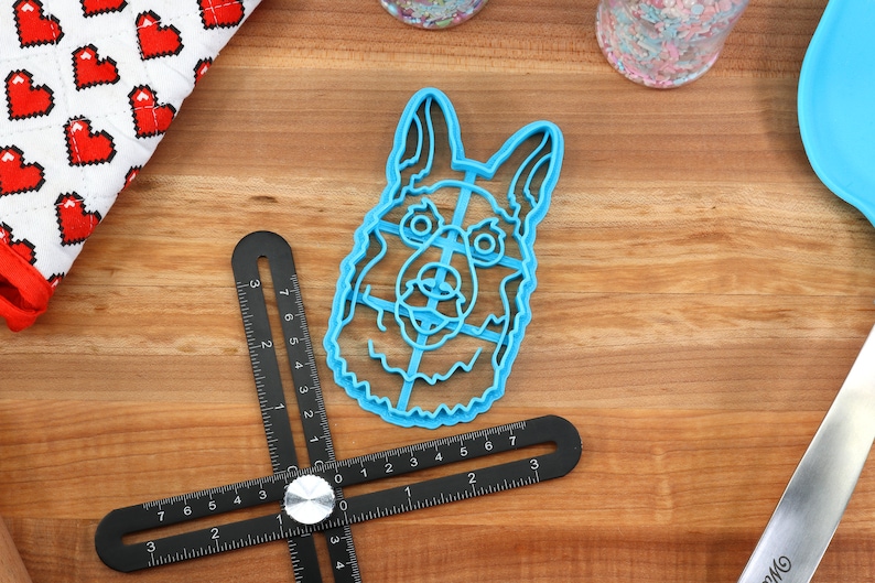 May include: A blue plastic cookie cutter shaped like a German Shepherd dog's head. The cutter is on a wooden surface with a ruler and a metal spatula.