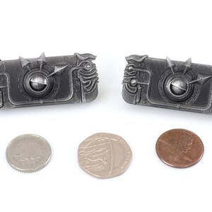 May include: Two silver-coloured metal keychains shaped like a rectangular box with intricate details and a small round knob in the centre. The keychains are about the size of a UK penny.