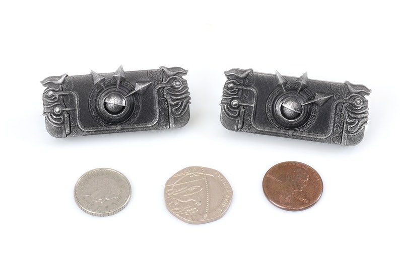 May include: Two silver-coloured metal keychains shaped like a rectangular box with intricate details and a small round knob in the centre. The keychains are about the size of a UK penny.