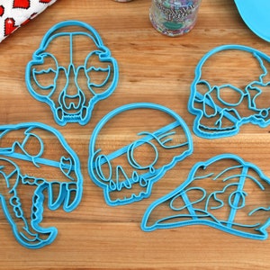 May include: Five blue plastic cookie cutters shaped like animal skulls. The cutters are on a wooden surface.