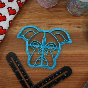 May include: A blue plastic cookie cutter shaped like a boxer dog's face. The cutter has a detailed design with a nose, eyes, and ears.