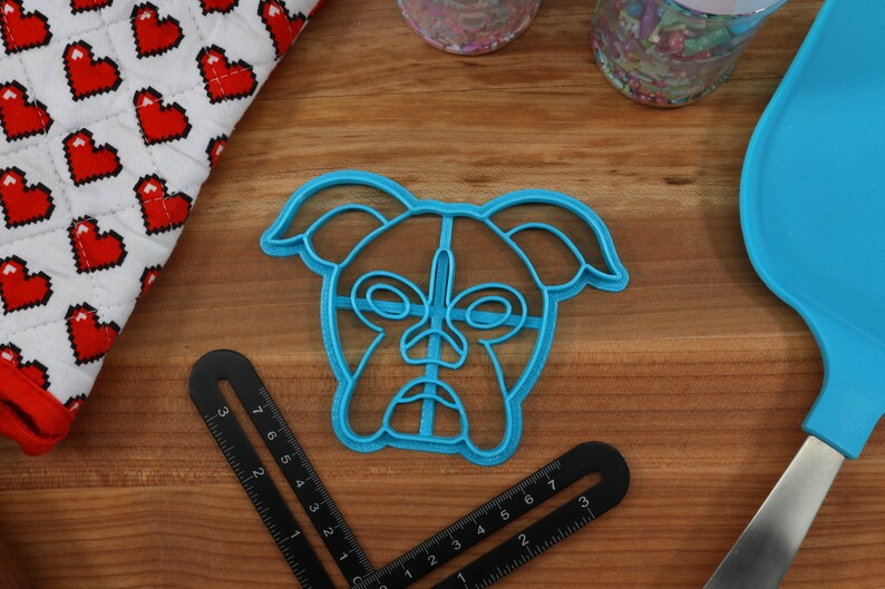 May include: A blue plastic cookie cutter shaped like a boxer dog's face. The cutter has a detailed design with a nose, eyes, and ears.