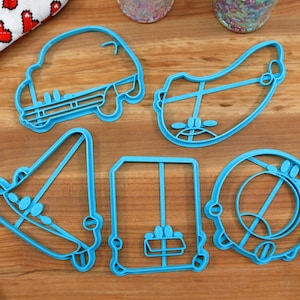 Könnte beinhalten: Fünf blaue Plastik-Ausstecher in Form eines Autos, eines Gesichts, eines Hutes, einer Schachtel und eines Kreises. Die Ausstecher liegen auf einer Holzoberfläche.