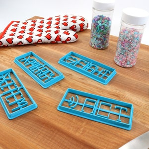 May include: Four blue plastic cookie cutters on a wooden surface. The cutters are shaped like the words "Merry", "Fight", "Off", and "Eat". There are two jars of sprinkles in the background.