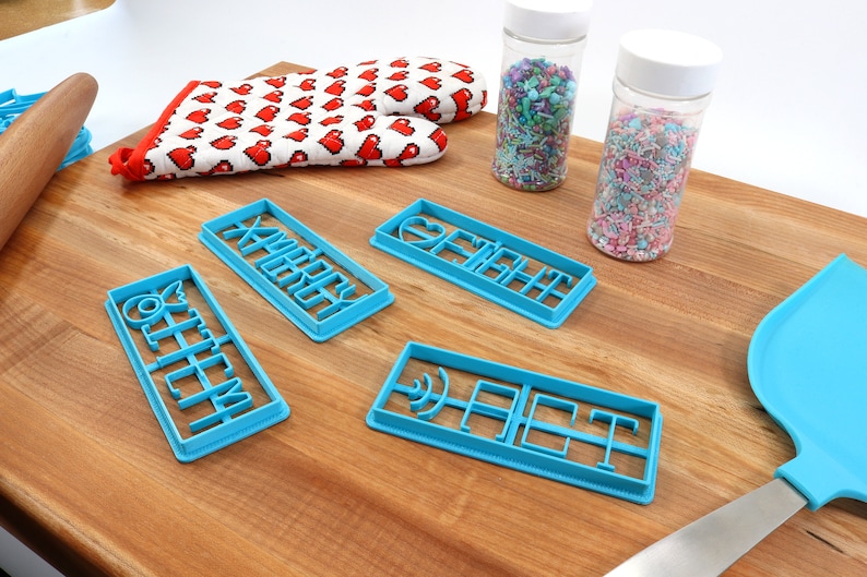 May include: Four blue plastic cookie cutters on a wooden surface. The cutters are shaped like the words "Merry", "Fight", "Off", and "Eat". There are two jars of sprinkles in the background.
