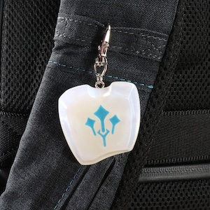 May include: A white, opaque charm with a blue symbol on a silver chain attached to a black backpack.