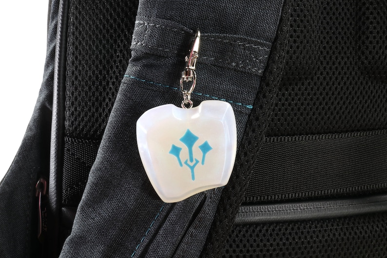 May include: A white, opaque charm with a blue symbol on a silver chain attached to a black backpack.