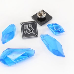 May include: A silver pin with a black background and a design of three swords. There are four blue crystal-like objects on a white background.